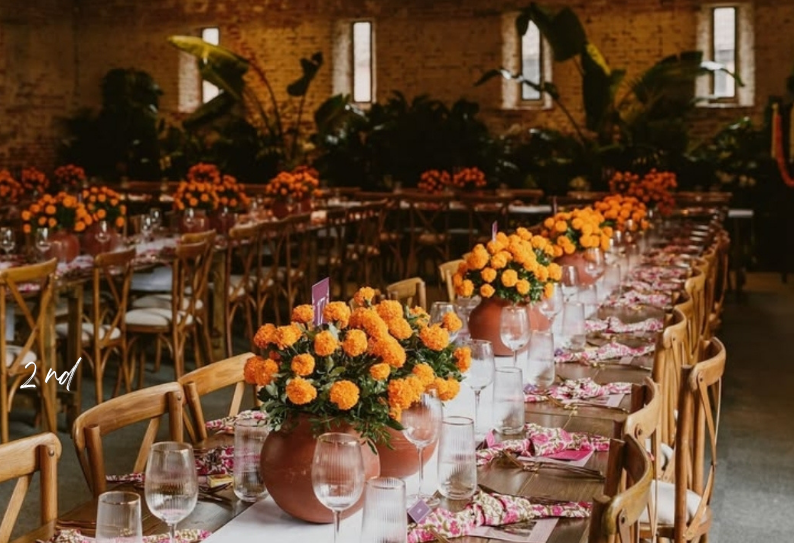 A long community dinner table set with orange marigold centerpieces, glassware, and warm brick surroundings.